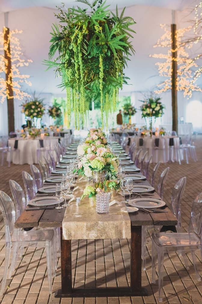 clear acrylic chairs under a long rectangular table with flowers down the center and green foliage baskets hanging from the ceiling
